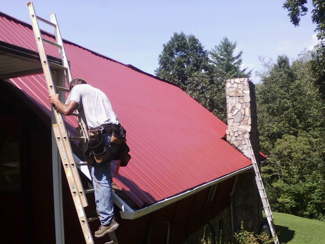 Roofer on a ladder beside a red metal roof and stone chimney — Soffit & Fascia Repair by Roofing Brothers in Kelford